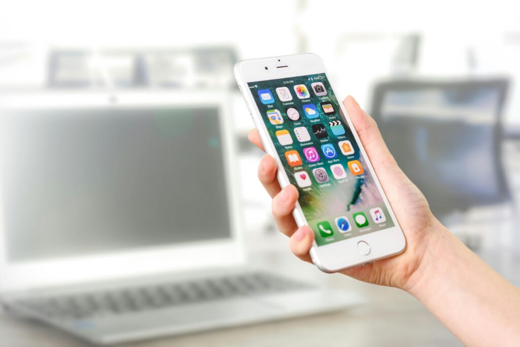 pexels-photo-887751-887751 Close-up view of a smartphone displaying apps, held by a hand, with a blurred laptop in the background.